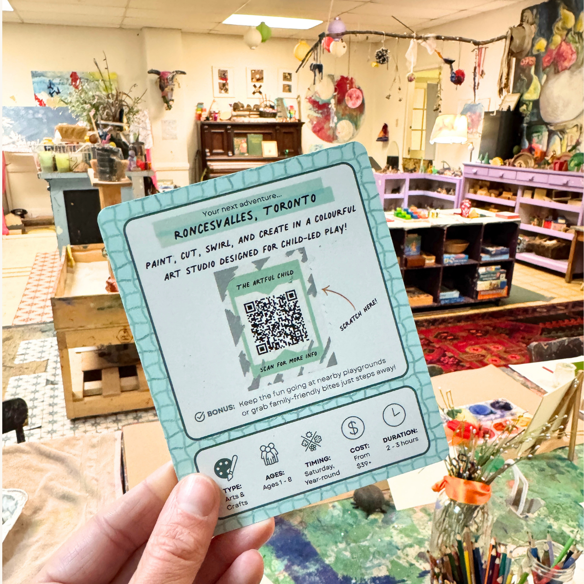 A hand holding a Roncesvalles card in a colourful kids' art studio.