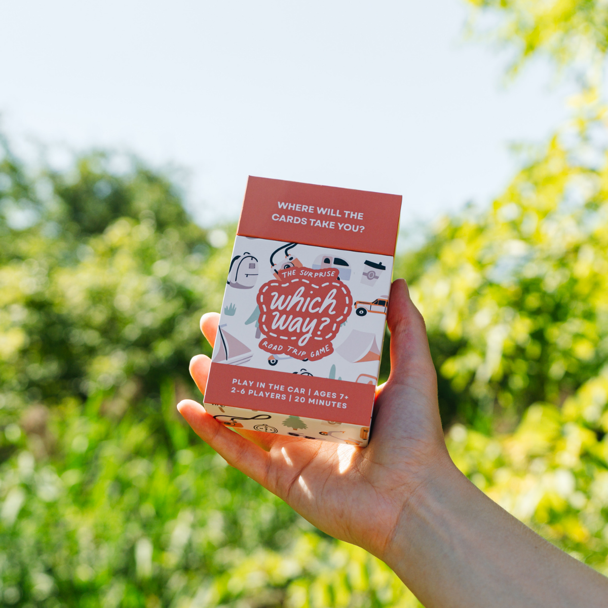 A hand holding a boxed card game outdoors against a blurred green garden background. The box is pink and white with illustrated graphics and readable text including 'which way?' and 'where will the cards take you?'.