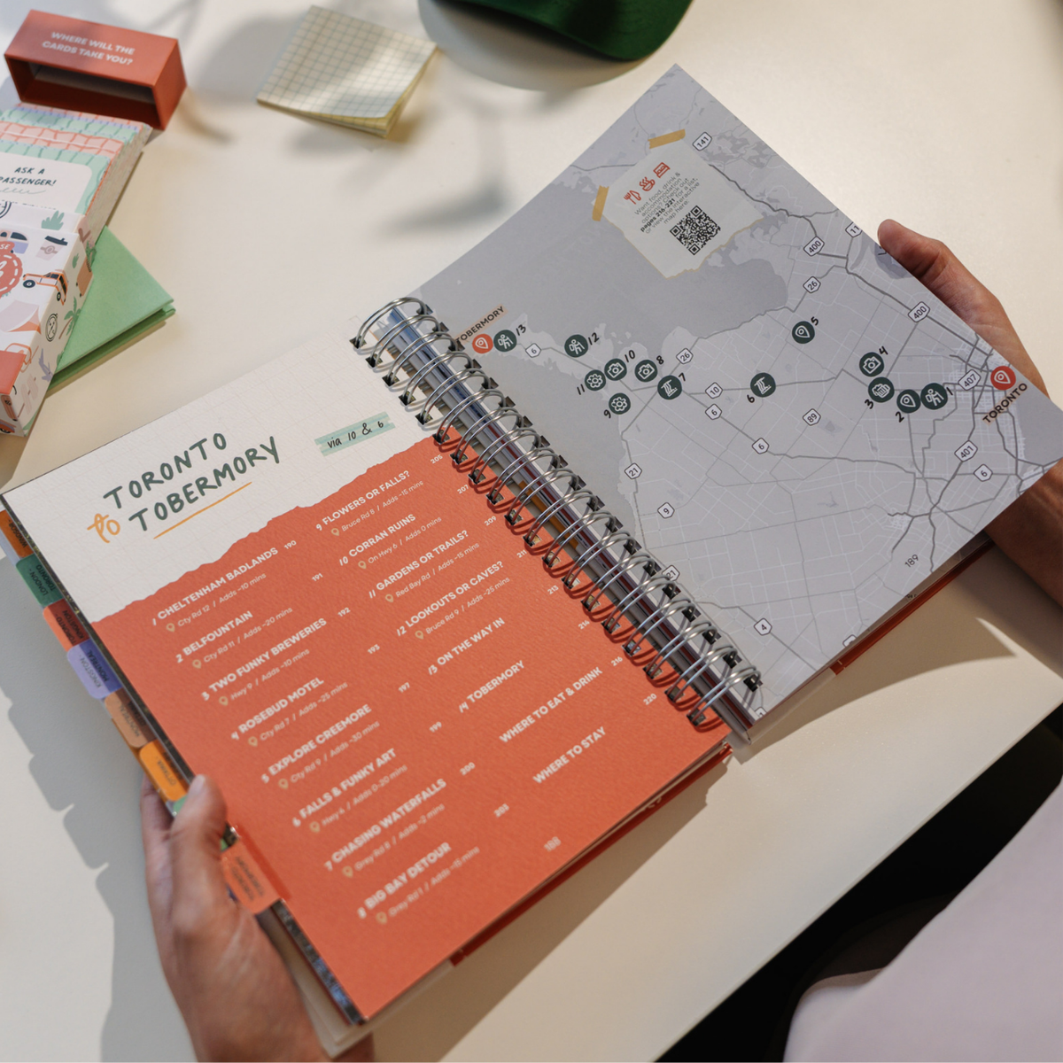 A person holds open a spiral-bound Toronto Tobermory travel planner on a light tabletop. The planner shows a map on the right page and a colorful itinerary list on the left page in orange, white, and coral tones.