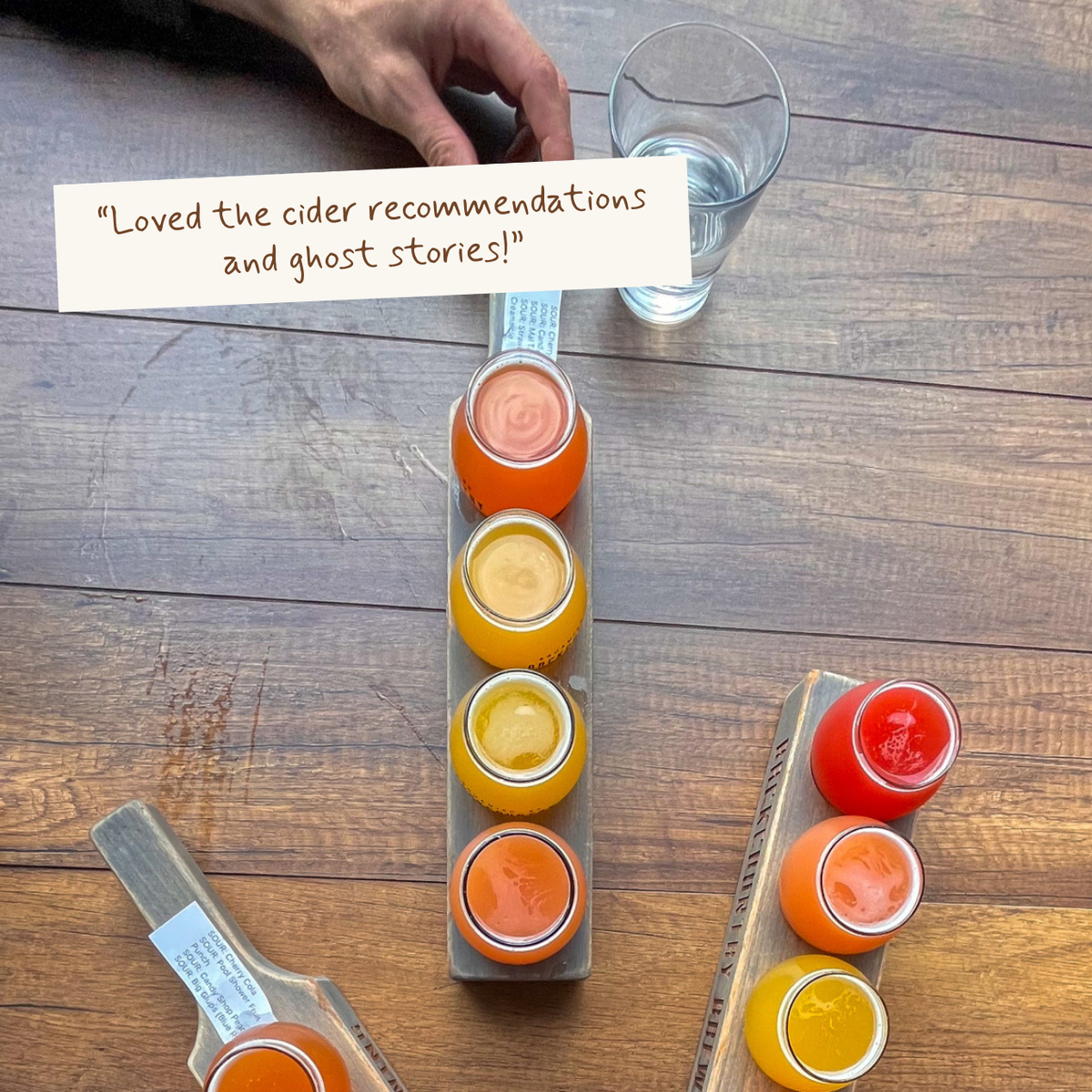 A top-down view of a wooden table with two cider flights, each holding four small glass jars of different colored cider. A hand is visible holding a glass, and a paper note with a customer review is placed on the table.