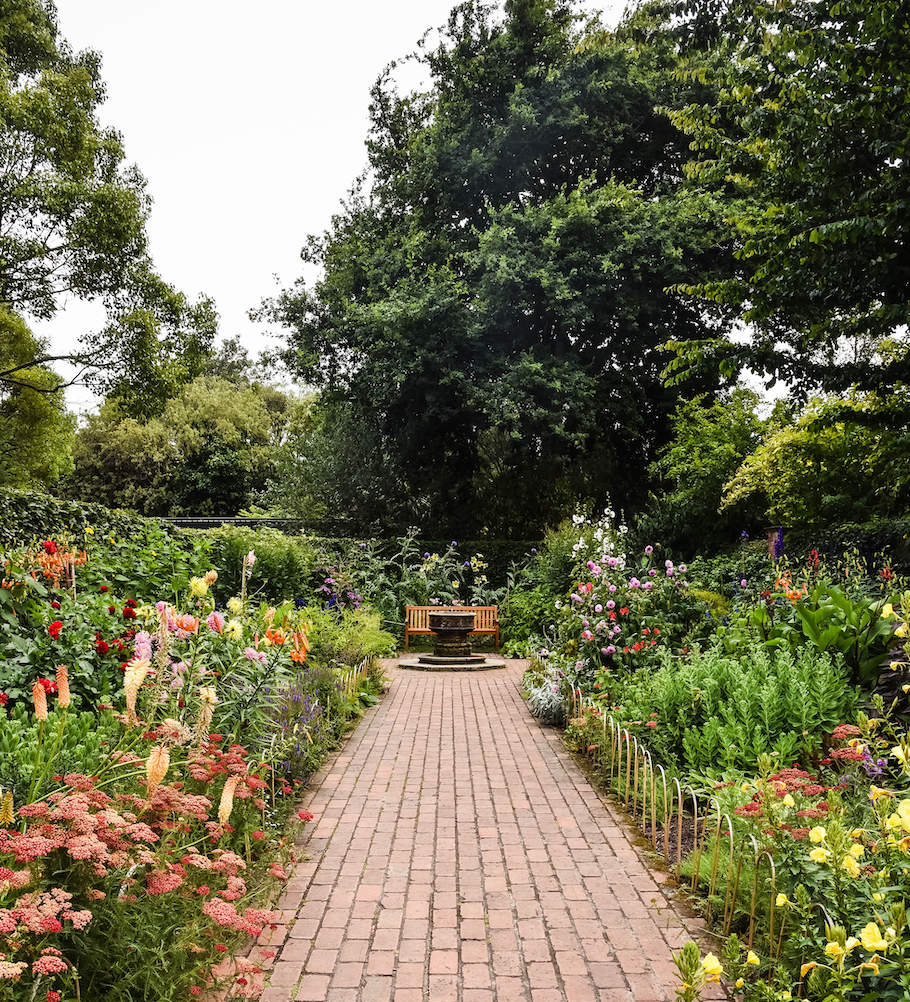 Park bench and fountain at the end of a brick path in a garden.