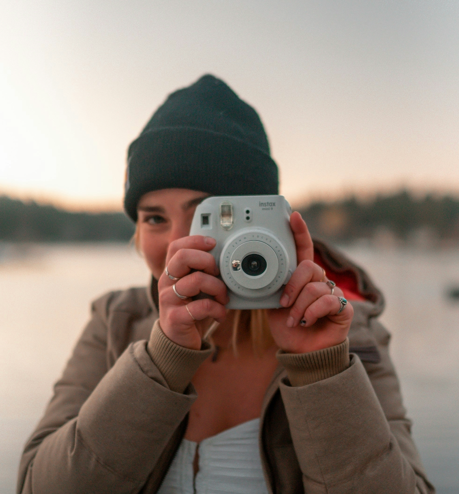 A person outdoors holding a camera up to their face, with a body of water and a blurred background.