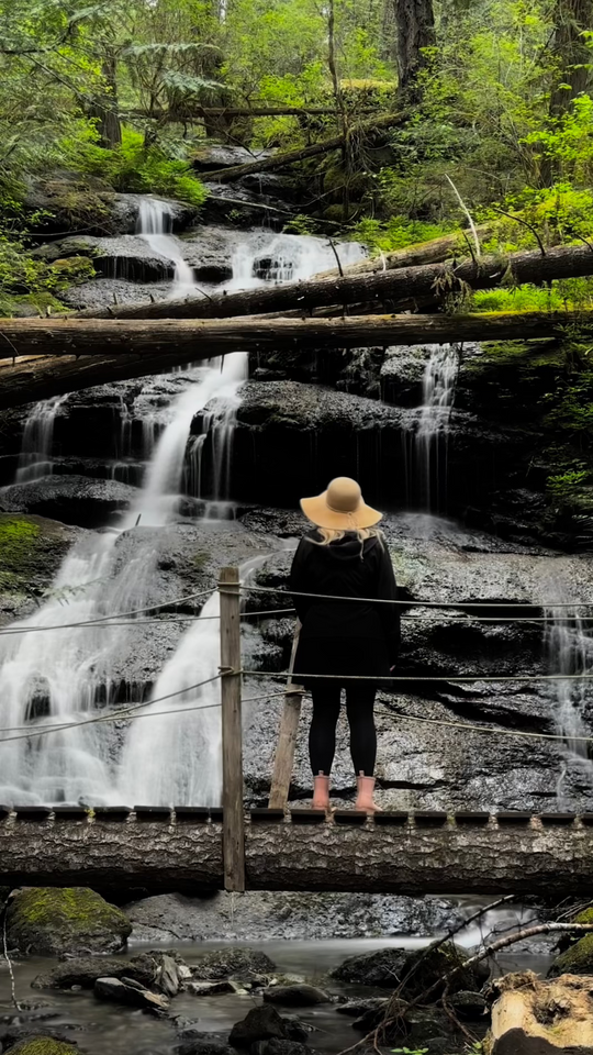 Woman with straw hat standing on bridge overlooking waterfall in a forest.