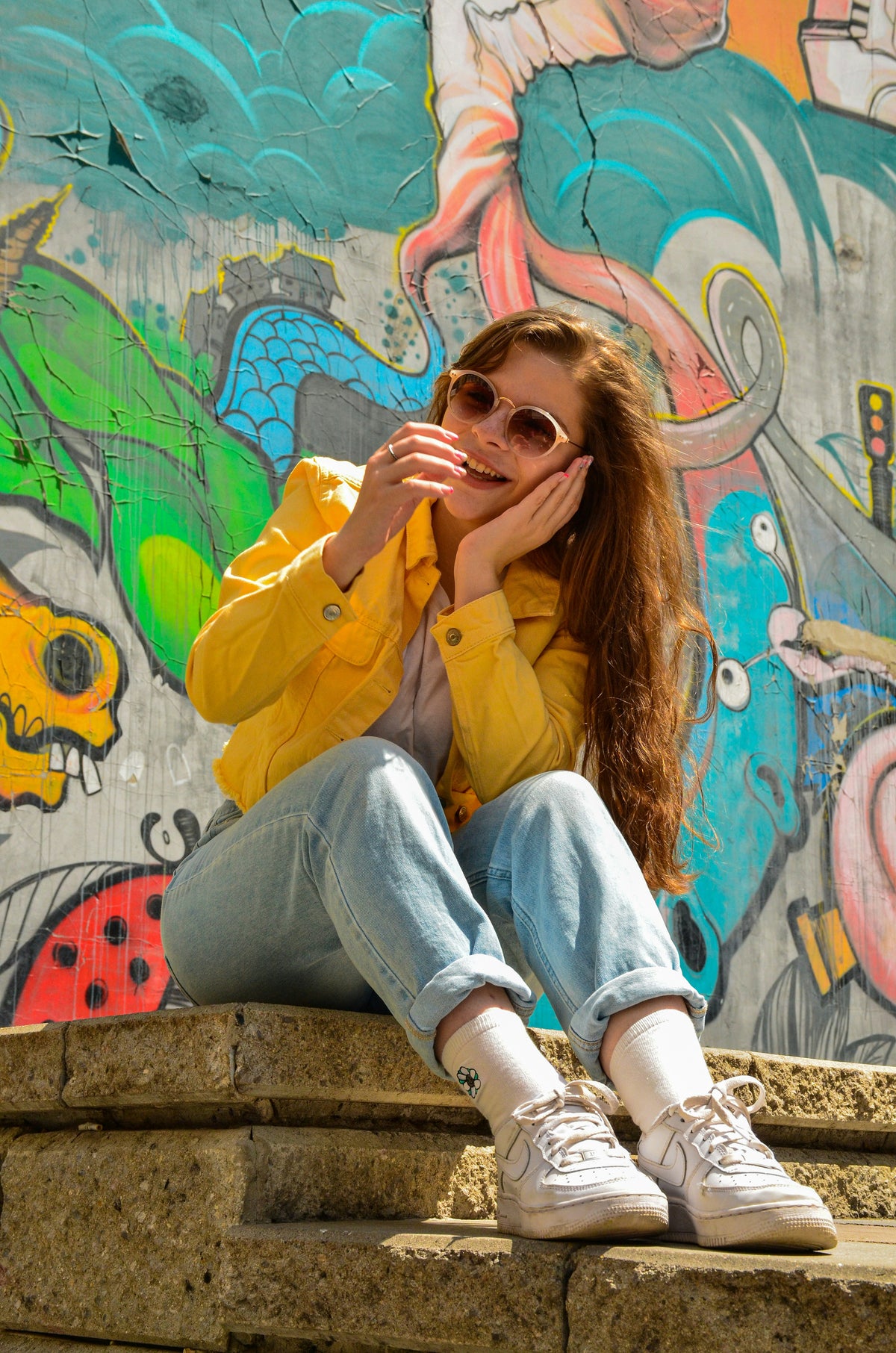 A woman sitting in front of a colourful mural, drinking a coffee.