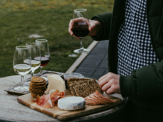 Person holding a glass of red wine with a charcuterie board and glasses of white wine on a wooden table outdoors.