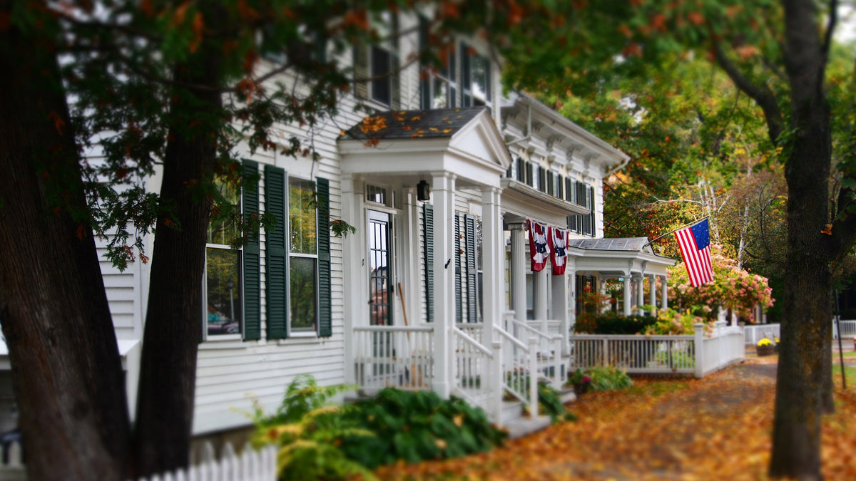 A white colonial house with an American flag and fall leaves.