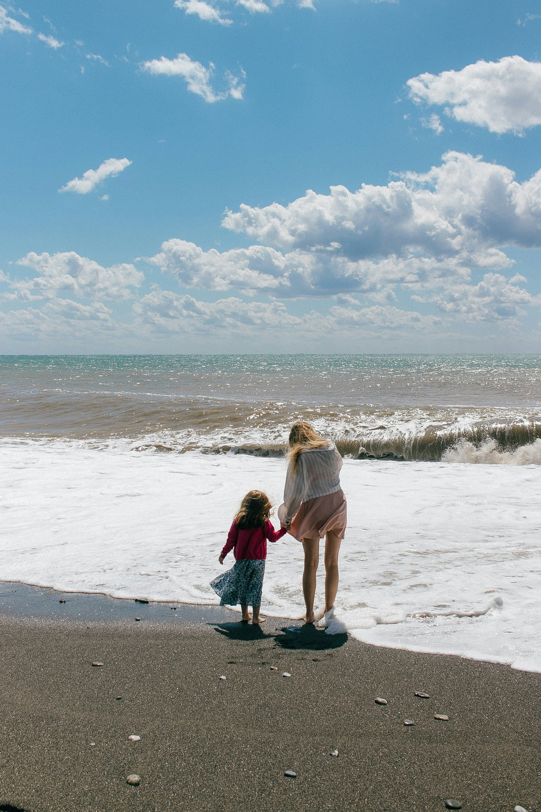 Woman holding hands with small child on the beach in front of the ocean.