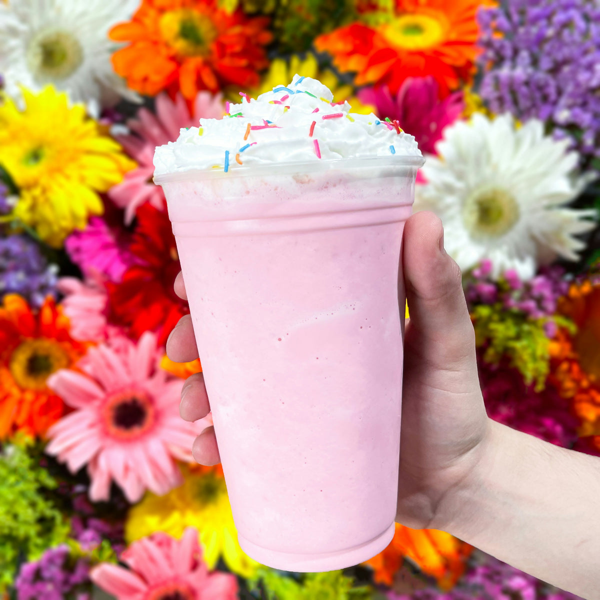 A pink milkshake held up against colourful flowers.