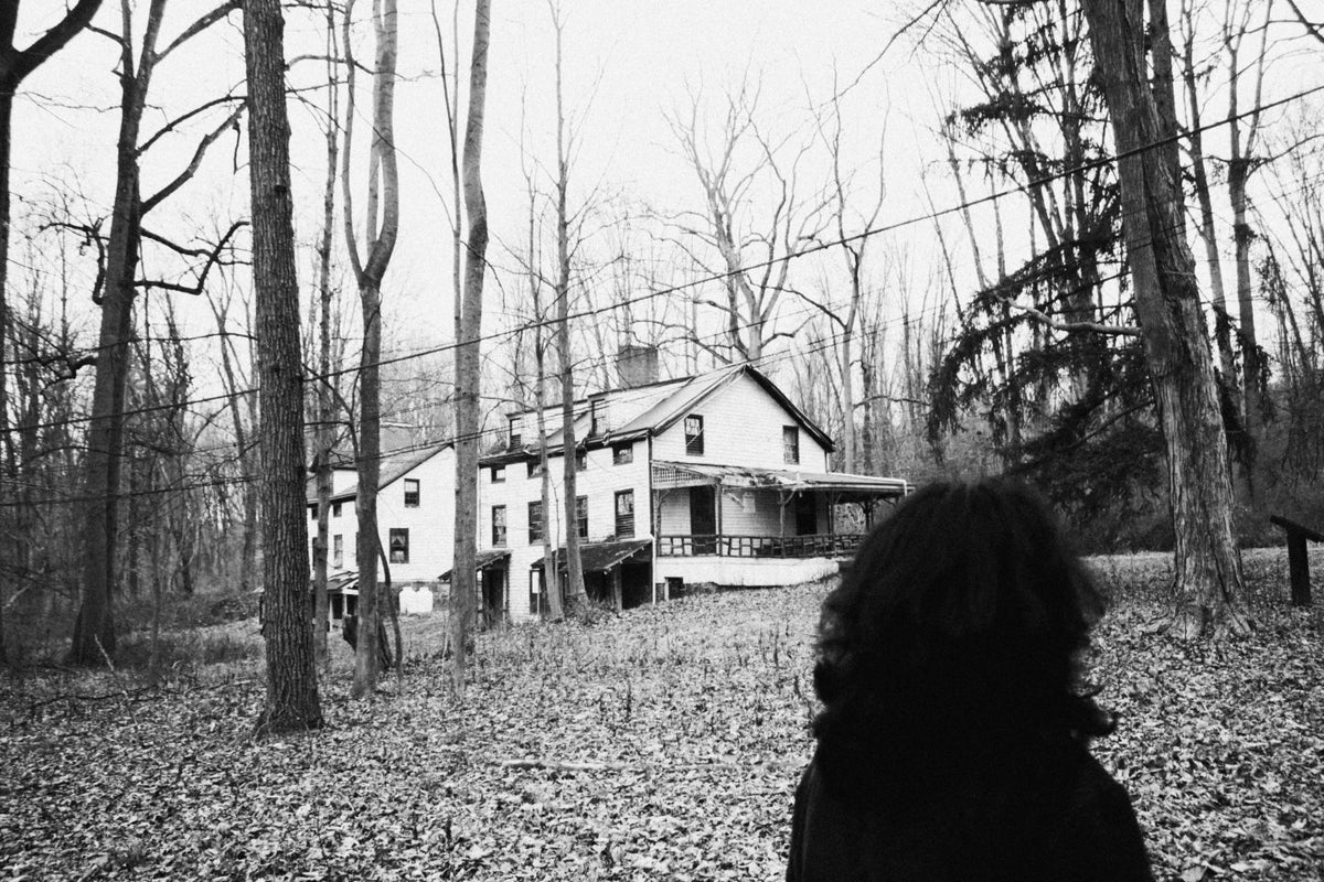 Black and white photo of a woman looking toward an old house through bare trees.