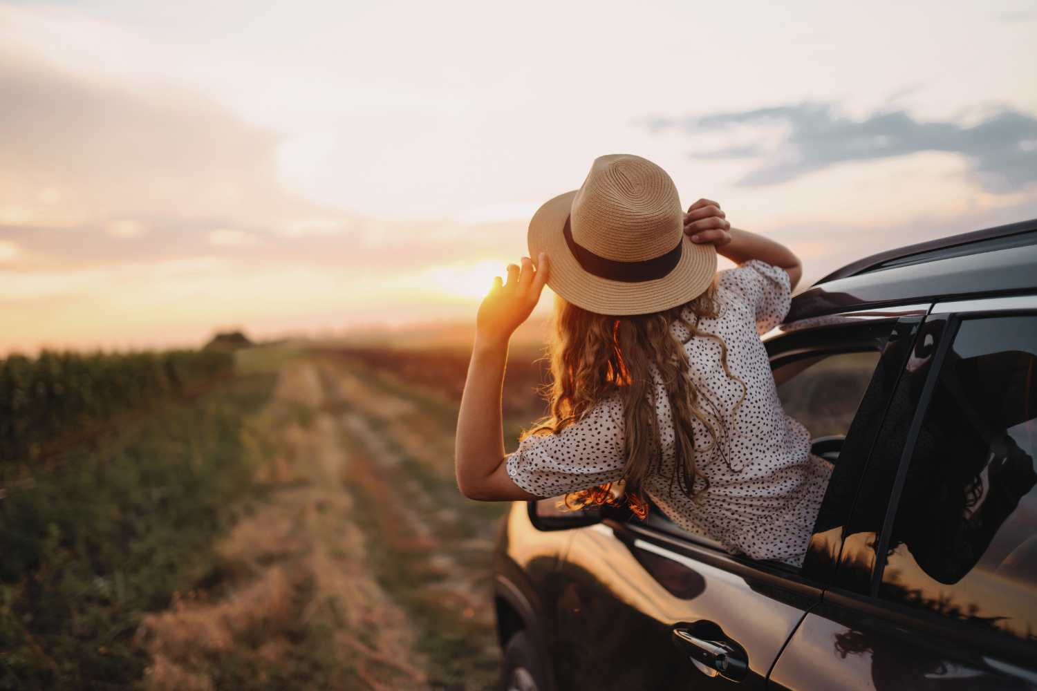 Woman with sunhat peering out of car window on dirt road at sunset.