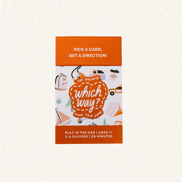 The Surprise Which Way? Road Trip Game box on a light cream textured background. The packaging features a bright orange top and bottom band with white text, and a central white panel decorated with cute illustrations of cars, a backpack, a coffee cup, and a suitcase, all framed by a dashed orange border.