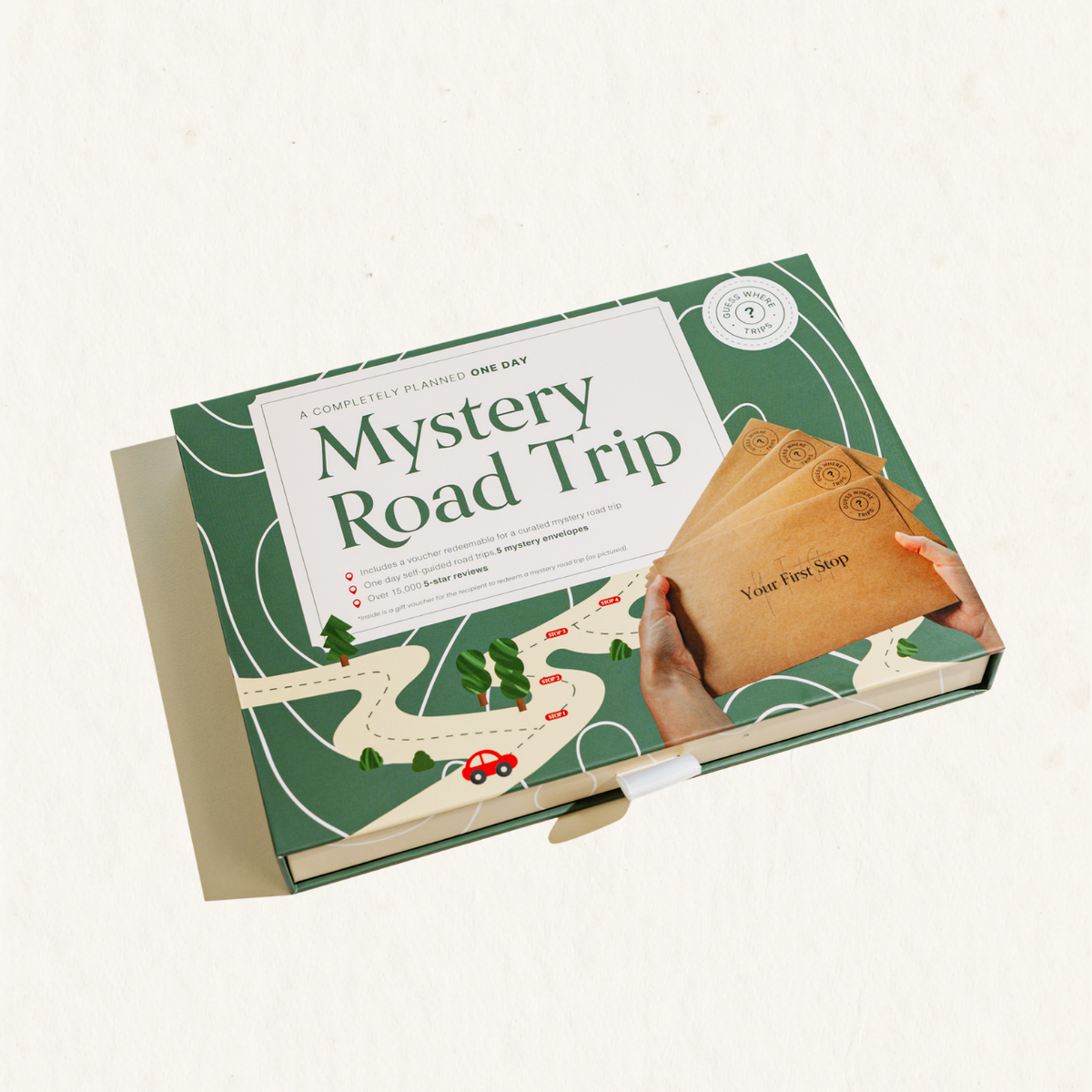 A boxed product labeled 'Mystery Road Trip' shown at an angle on a light cream background. The box is green with illustrated road and tree graphics, a large white title panel, and a photo of hands holding brown envelope-style cards.