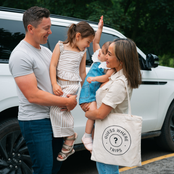 A family of four stands next to a white SUV in a parking lot. The mother holds a cream canvas tote bag with the text 'GUESS WHERE TRIPS?' and a question mark, while the father holds a girl in a striped dress and the mother holds a baby in a denim outfit.