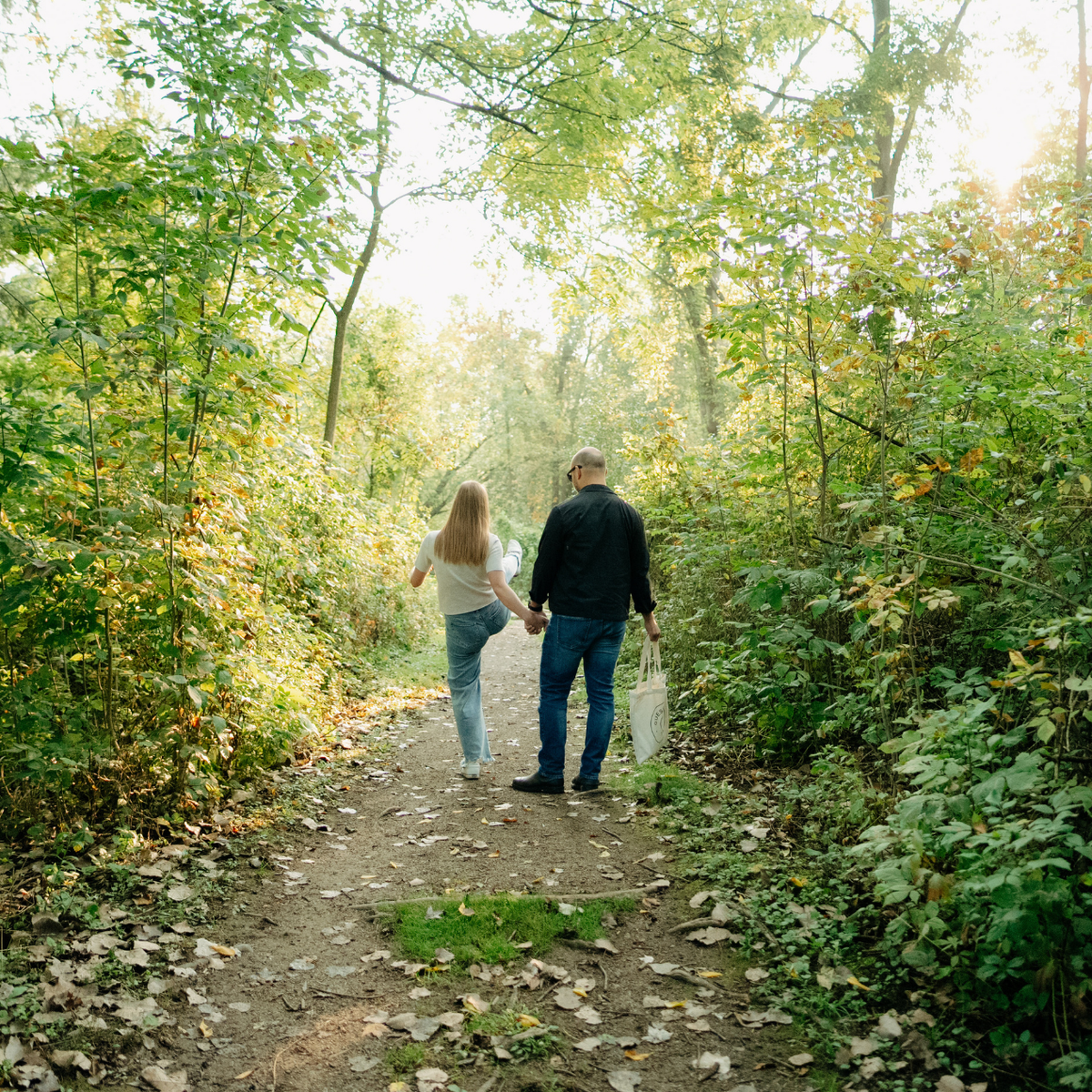 A couple holding hands walks away on a sunlit forest path surrounded by green trees and fallen leaves.