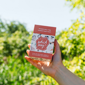 A hand holding a boxed card game outdoors against a blurred green garden background. The box is pink and white with illustrated graphics and readable text including 'which way?' and 'where will the cards take you?'.