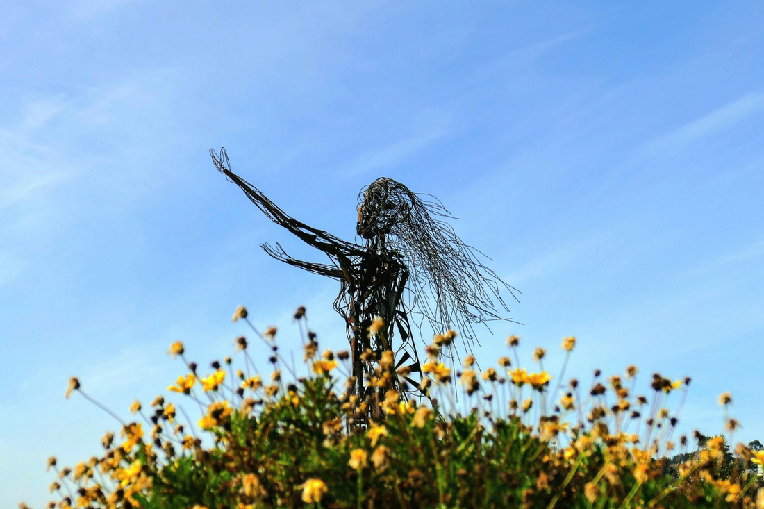 Sculpture of a woman with long hair in front of blue skies and behind a bush of yellow flowers.