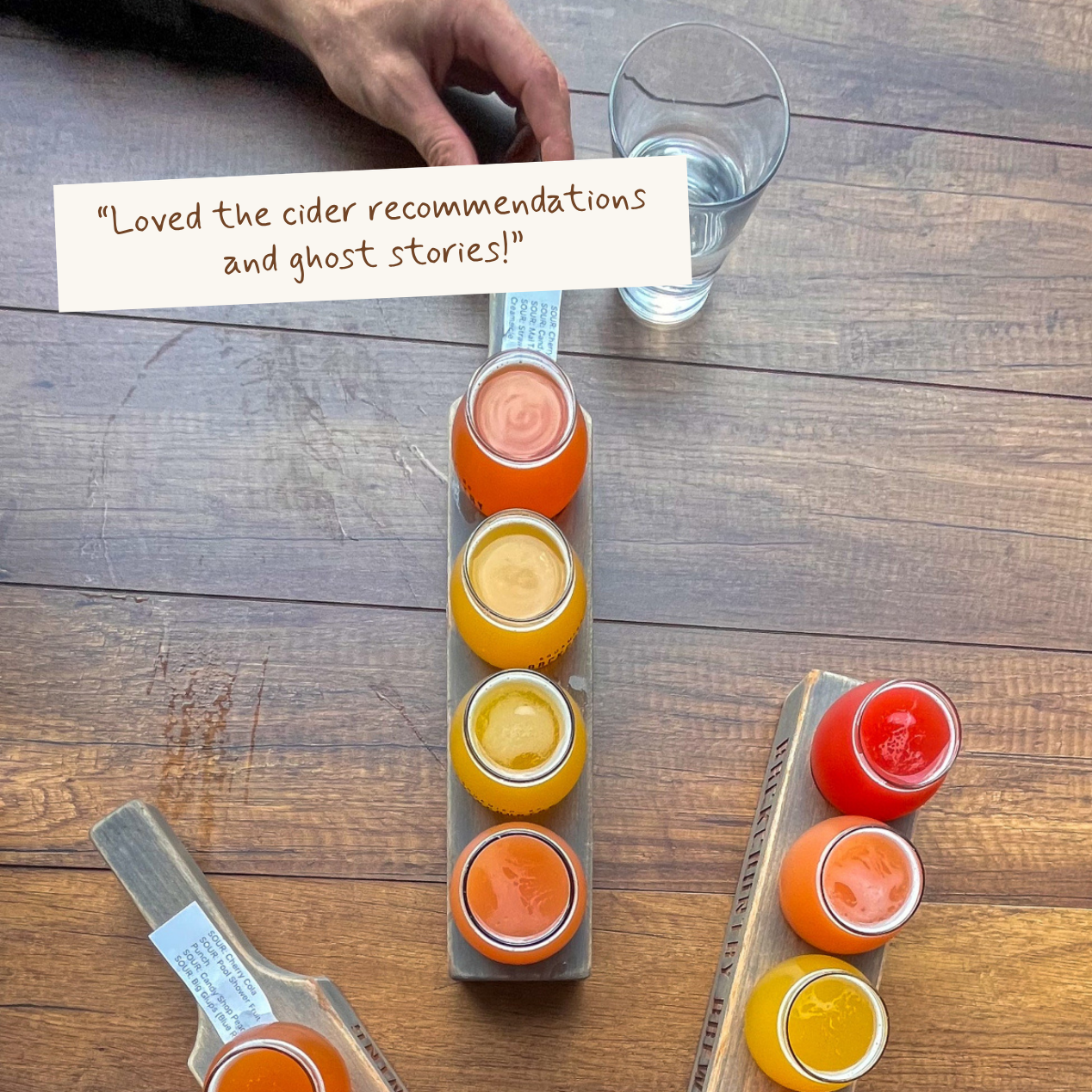 A top-down view of a wooden table with two cider flights, each holding four small glass jars of different colored cider. A hand is visible holding a glass, and a paper note with a customer review is placed on the table.