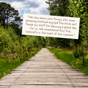 A handwritten note with brown ink is shown on a white background, superimposed over a scenic photograph of a wooden boardwalk path winding through a lush green forest under a cloudy sky.