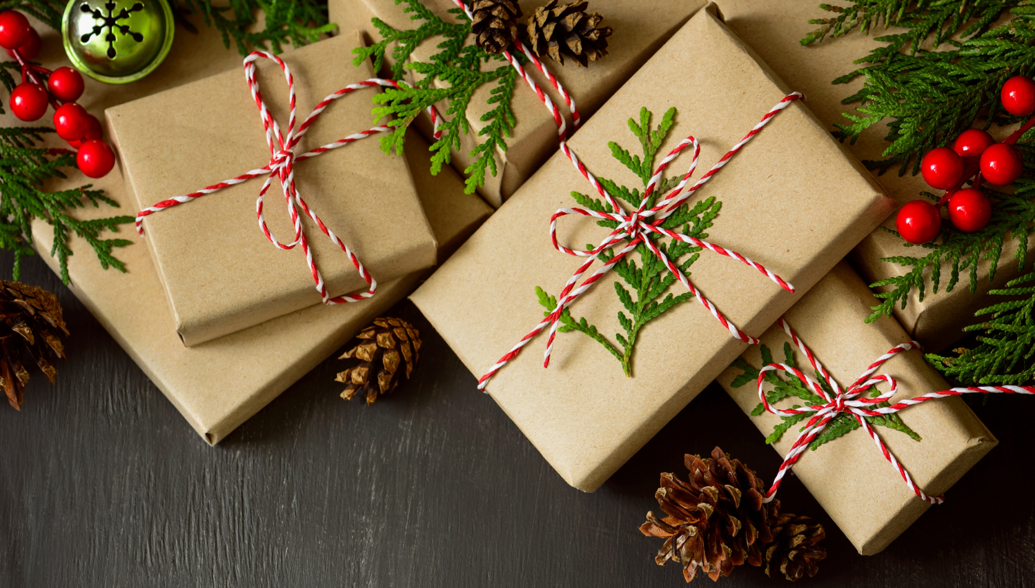 Gift boxes wrapped in brown paper with string and greenery, surrounded by pine cones and berries on a dark surface.