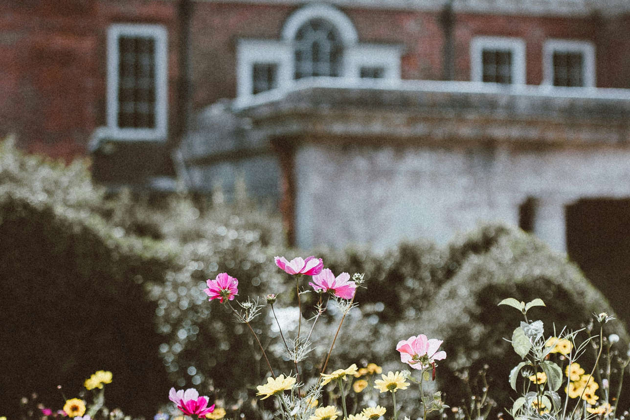 A photo depicting a garden with colorful flowers in the foreground, with a blurred background showing a large, elegant house.