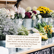 A rustic market stall filled with metal buckets of flowers, including white tulips, yellow daisies, burgundy chrysanthemums, and green foliage. A cream-colored paper note with handwritten brown text is placed in the foreground.