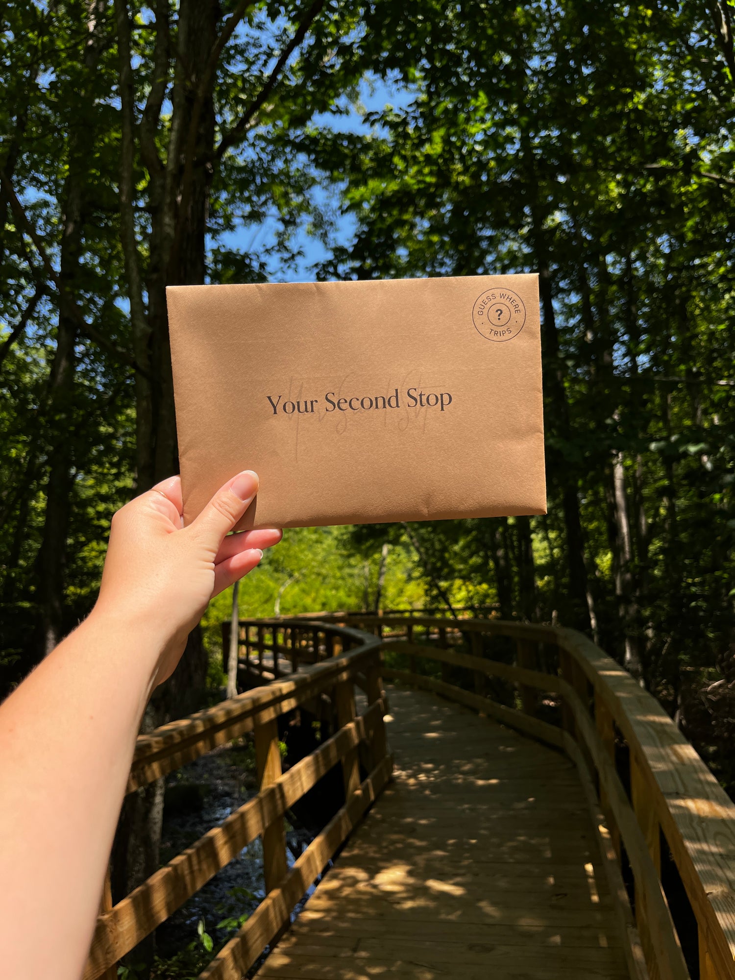 Arm holding mystery road trip envelope in front of trees and boardwalk