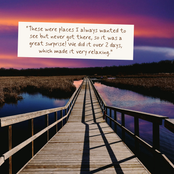 A scenic photograph of a wooden boardwalk leading over calm water toward a distant dock, with a purple and pink sunset sky above and a white text box overlaid near the top containing a quote.