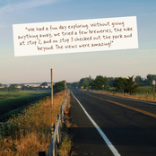 A scenic road stretches through a rural landscape under a pale blue sky, with a white paper note overlaid near the top containing a handwritten-style quote in brown text.