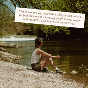 A woman sits on the shore of a calm river, looking out at the water, with a white text overlay in the upper right that reads, 'T"he itinerary was incredibly well planned, with a perfect balance of charming small towns, unique local markets, and beautiful scenic stops.'.