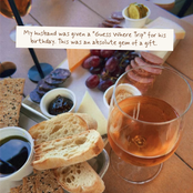 A wooden table styled with a charcuterie board and a glass of rosé wine, featuring sliced bread, crackers, cheese, grapes, salami, and small condiment cups. A white text overlay reads: 'My husband was given a "Guess Where Trip" for his birthday.