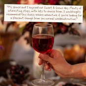 A hand holds a wine glass filled with red wine in a blurred outdoor setting, with a white text overlay that reads: 'My spouse and I enjoyed our Sweet & Scenic day. Plenty of interesting stops, with lots to choose from.