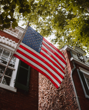A large American flag hanging on the side of a red brick building with greenery in the background.
