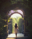 A person standing at the entrance of a lush garden pathway, looking towards the light at the end of the tunnel created by the archway.