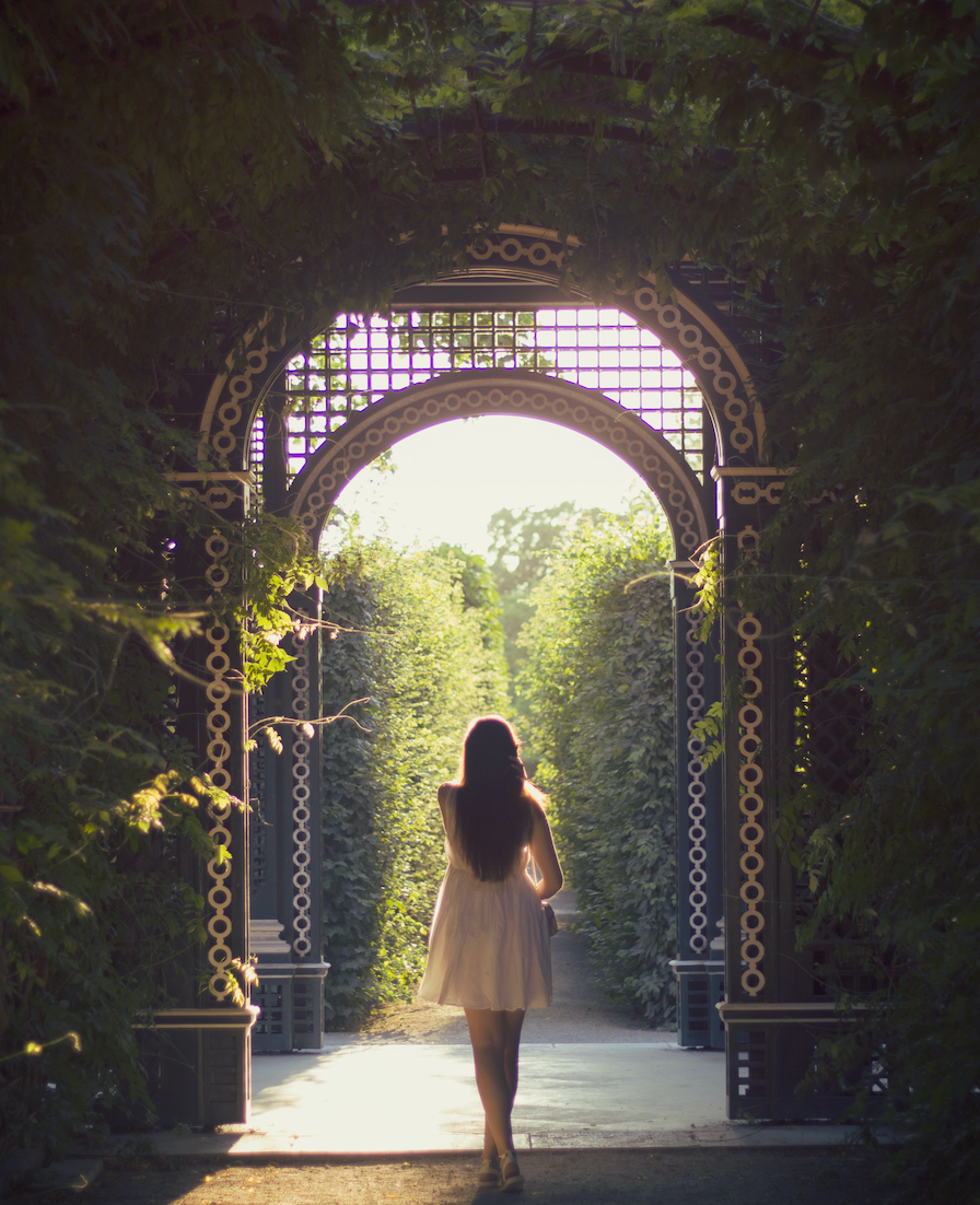 A person standing at the entrance of a lush garden pathway, looking towards the light at the end of the tunnel created by the archway.