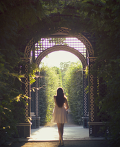 A person standing at the entrance of a lush garden pathway, looking towards the light at the end of the tunnel created by the archway.