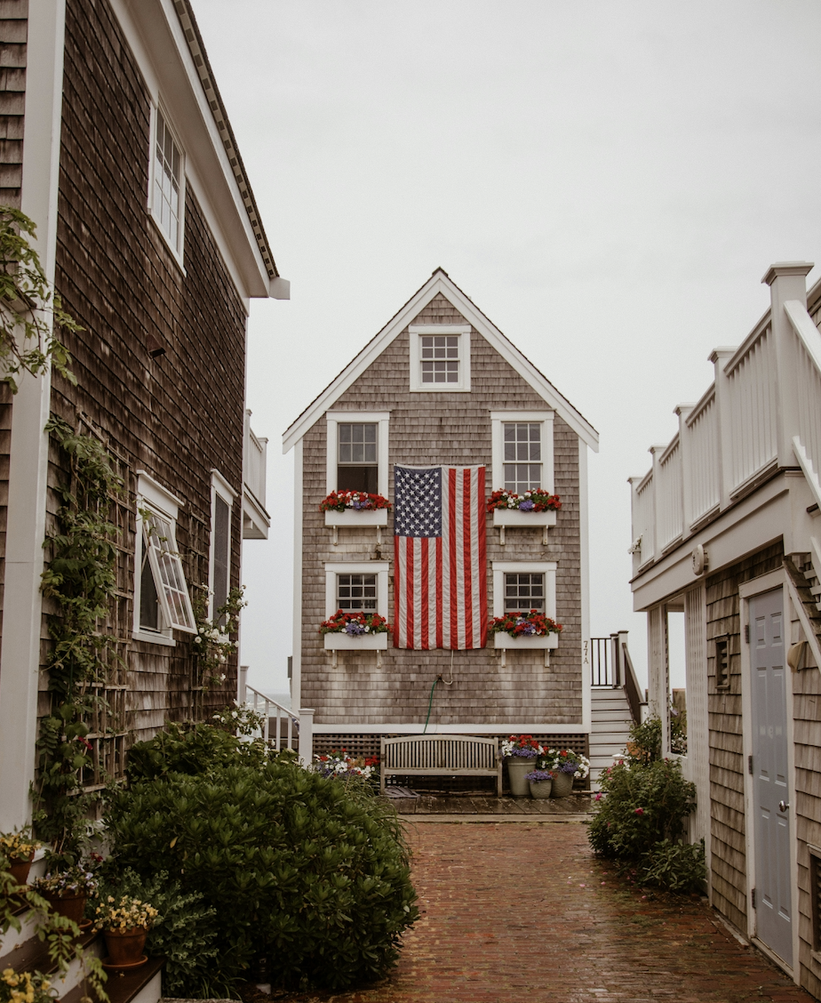 A picturesque street in a New England town with a two-story house adorned with an American flag, surrounded by cozy buildings and greenery.