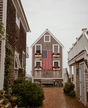 A picturesque street in a New England town with a two-story house adorned with an American flag, surrounded by cozy buildings and greenery.