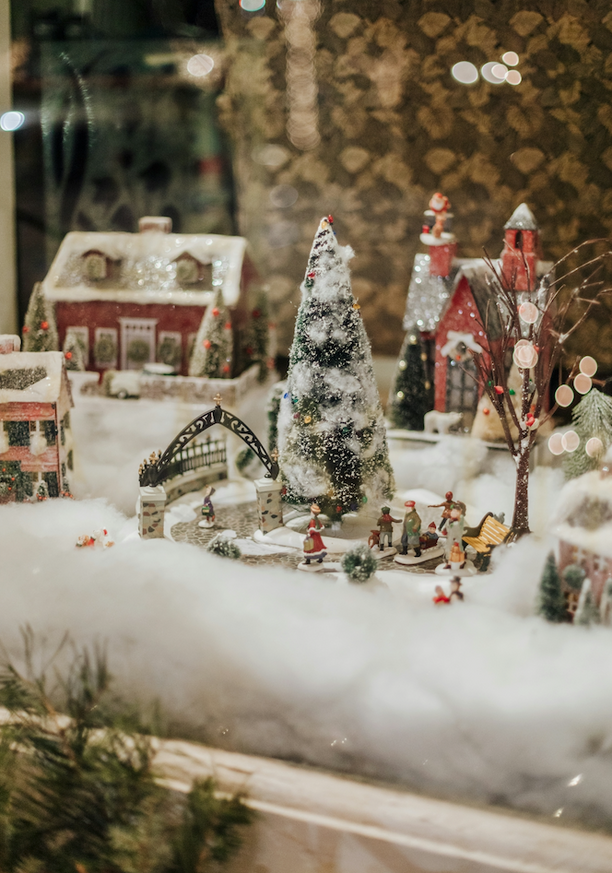 A festive Christmas-themed miniature village display with snow-covered houses and a large Christmas tree in the center.