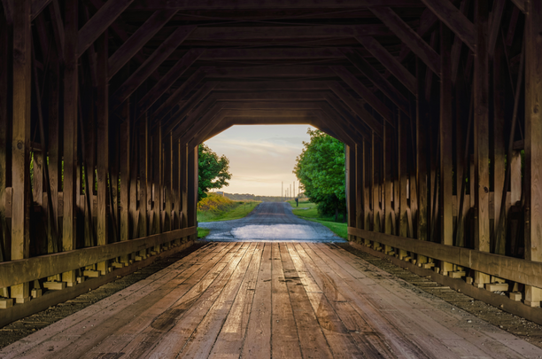 View from inside a wooden covered bridge looking out at a tree-lined road leading to a bright sky.