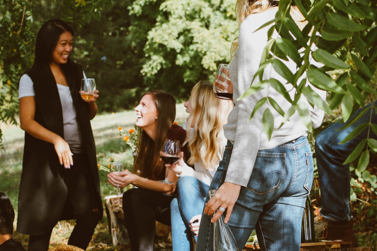 A group of people enjoying outdoor drinks and food, engaged in conversation in a green, leafy setting.
