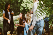 A group of people enjoying outdoor drinks and food, engaged in conversation in a green, leafy setting.