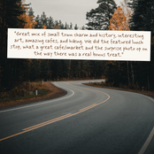 A winding asphalt road curves through a forested area with trees and autumn foliage in the background. A white rectangular text box is overlaid near the top center with a customer review quote in brown lettering.