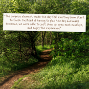 A wooden sign with a quote is placed on a dirt path in a lush green forest. The quote reads: 'The surprise element made the day feel exciting from start to finish.
