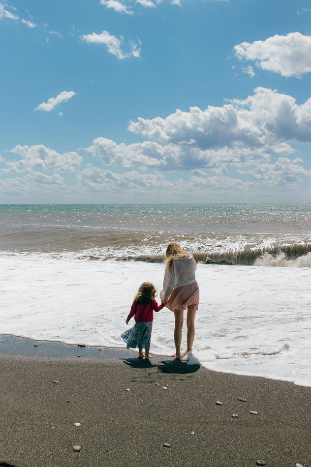Woman holding hands with small child on the beach in front of the ocean.