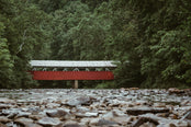 A red covered bridge crossing a rocky stream, surrounded by green forest.