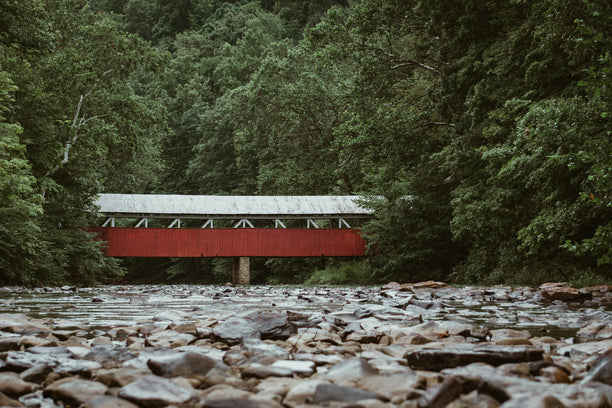 A red covered bridge crossing a rocky stream, surrounded by green forest.
