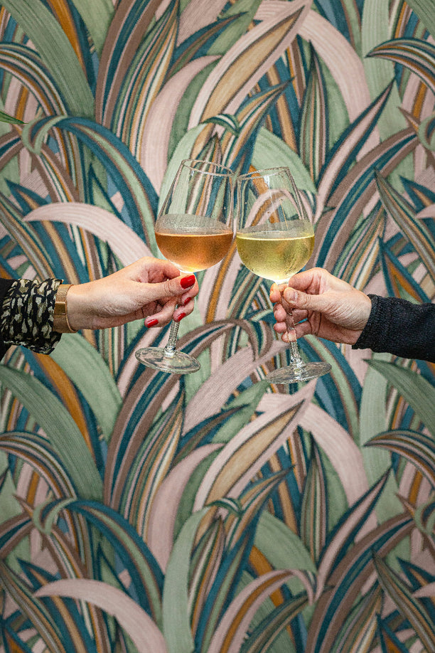Two people toasting with glasses of wine, set against a wallpaper with a tropical leaf pattern.