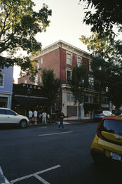 A street scene in a city with trees, buildings, and cars. People are walking on the sidewalk.