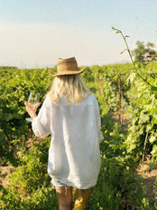Blonde woman walking with a glass of wine through a vineyard.