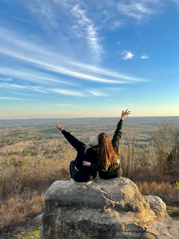 Two people sitting on a rock with arms raised, enjoying a scenic view of a landscape.